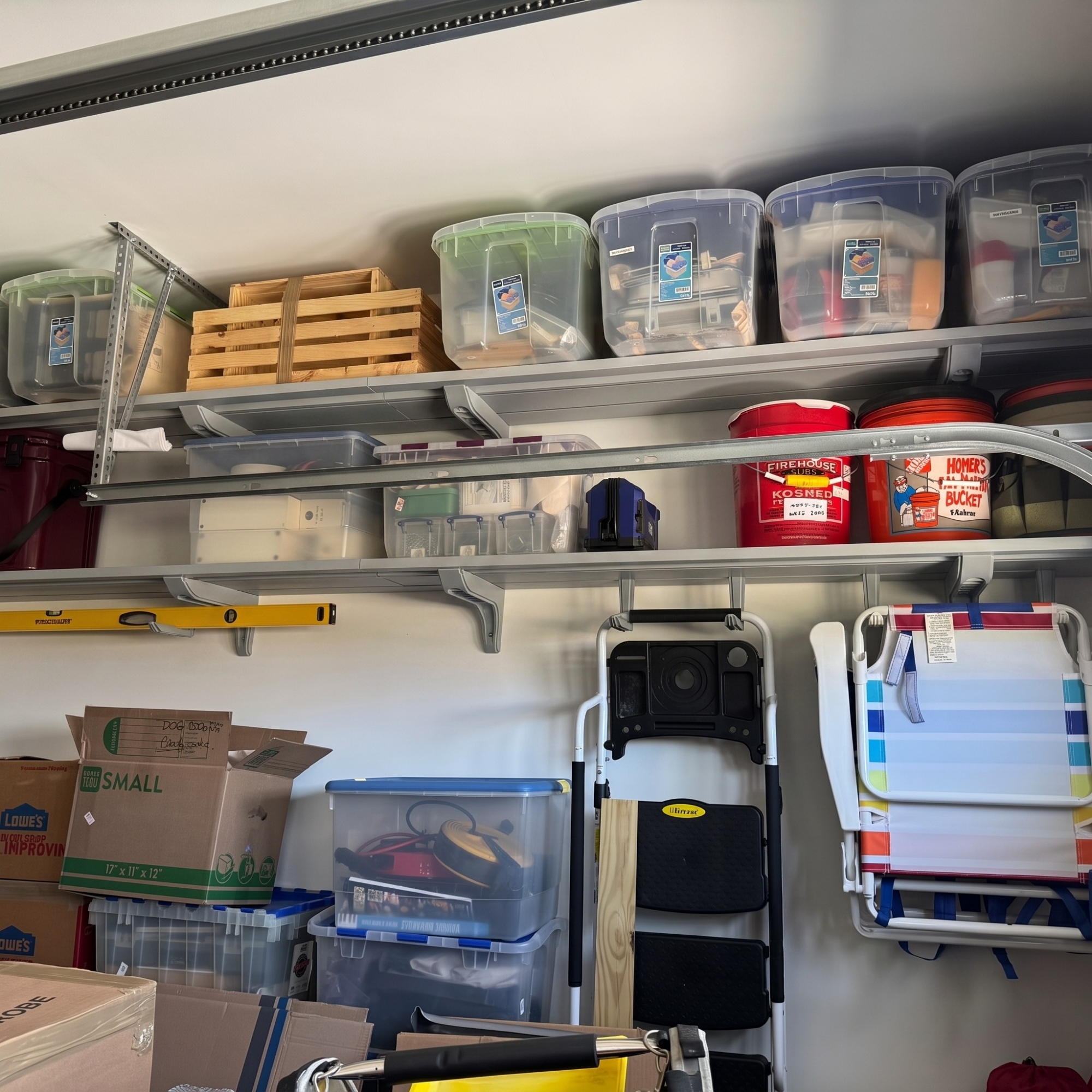 A garage storage setup with metal shelves holding clear plastic bins, wooden crates, and red buckets, alongside a folding step ladder and a wall-mounted organizer rack with papers.