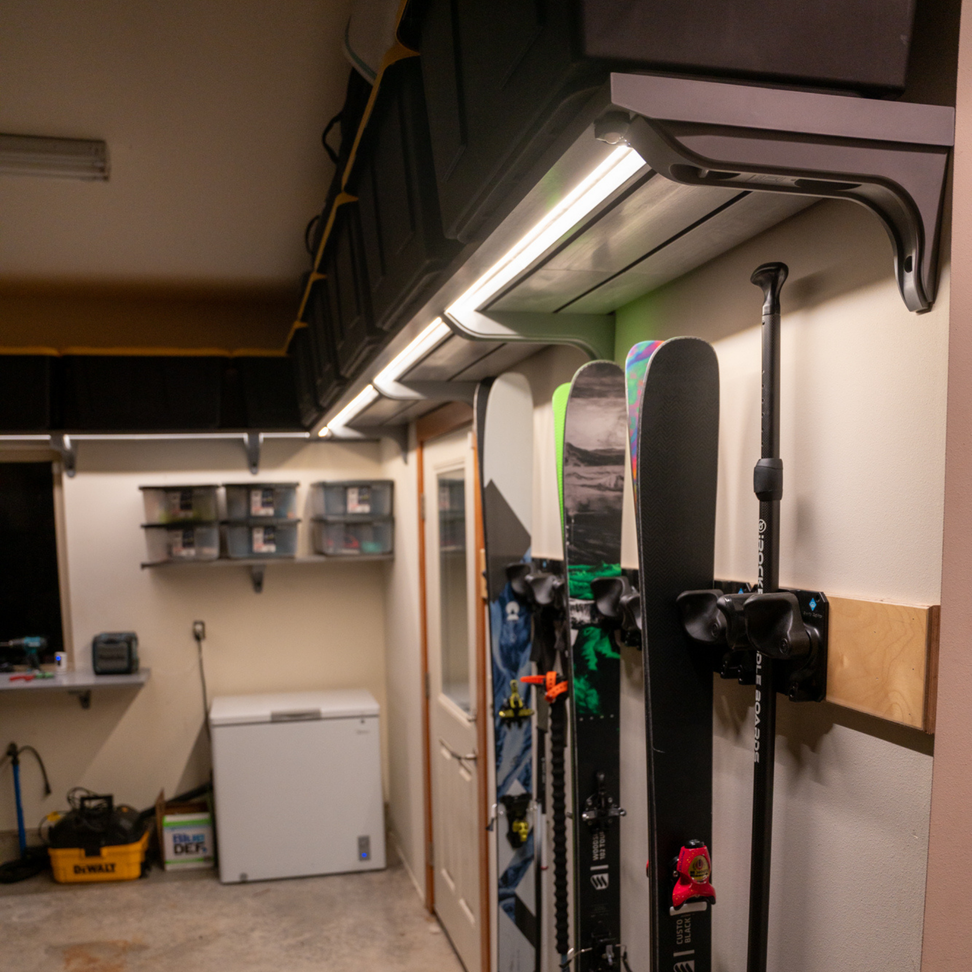 A ski rack mounted on a garage wall holding skis and ski poles, with storage bins and a chest freezer visible in the garage interior.
