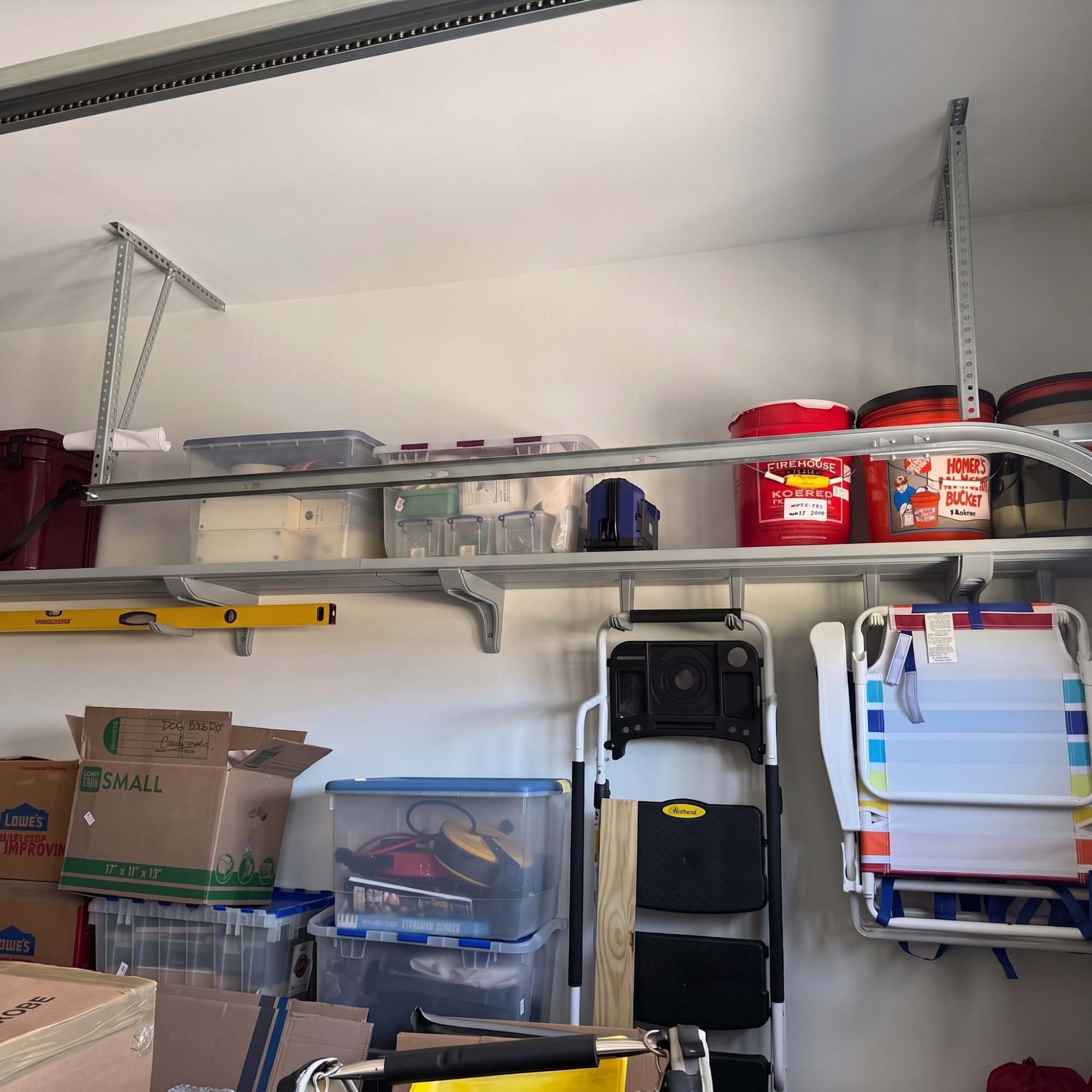 A garage storage setup with metal wall shelves holding plastic bins, buckets, a folding chair, and boxes against a white wall.