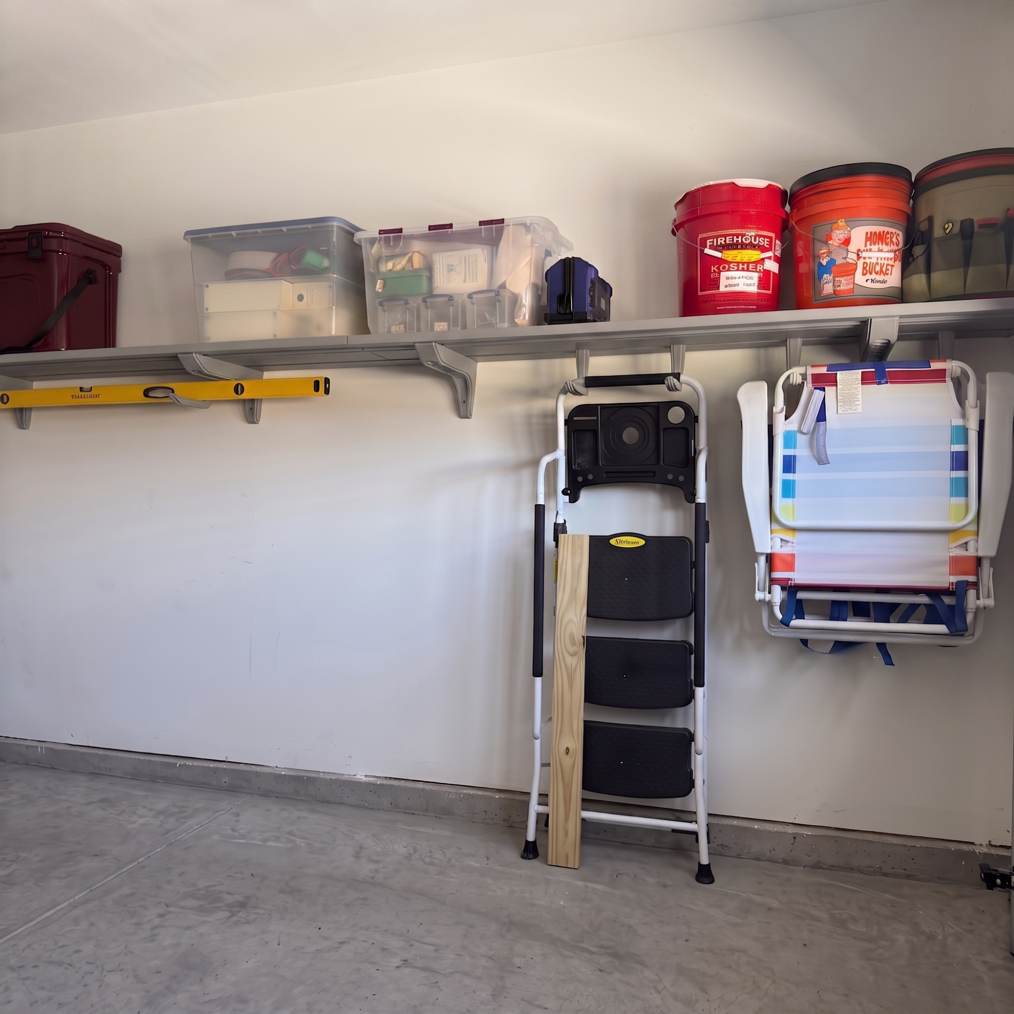 A metal garage wall-mounted storage shelf system holding various household items, including plastic bins, buckets, a yellow level, a folding step ladder, and a white folding chair, against a white garage wall.