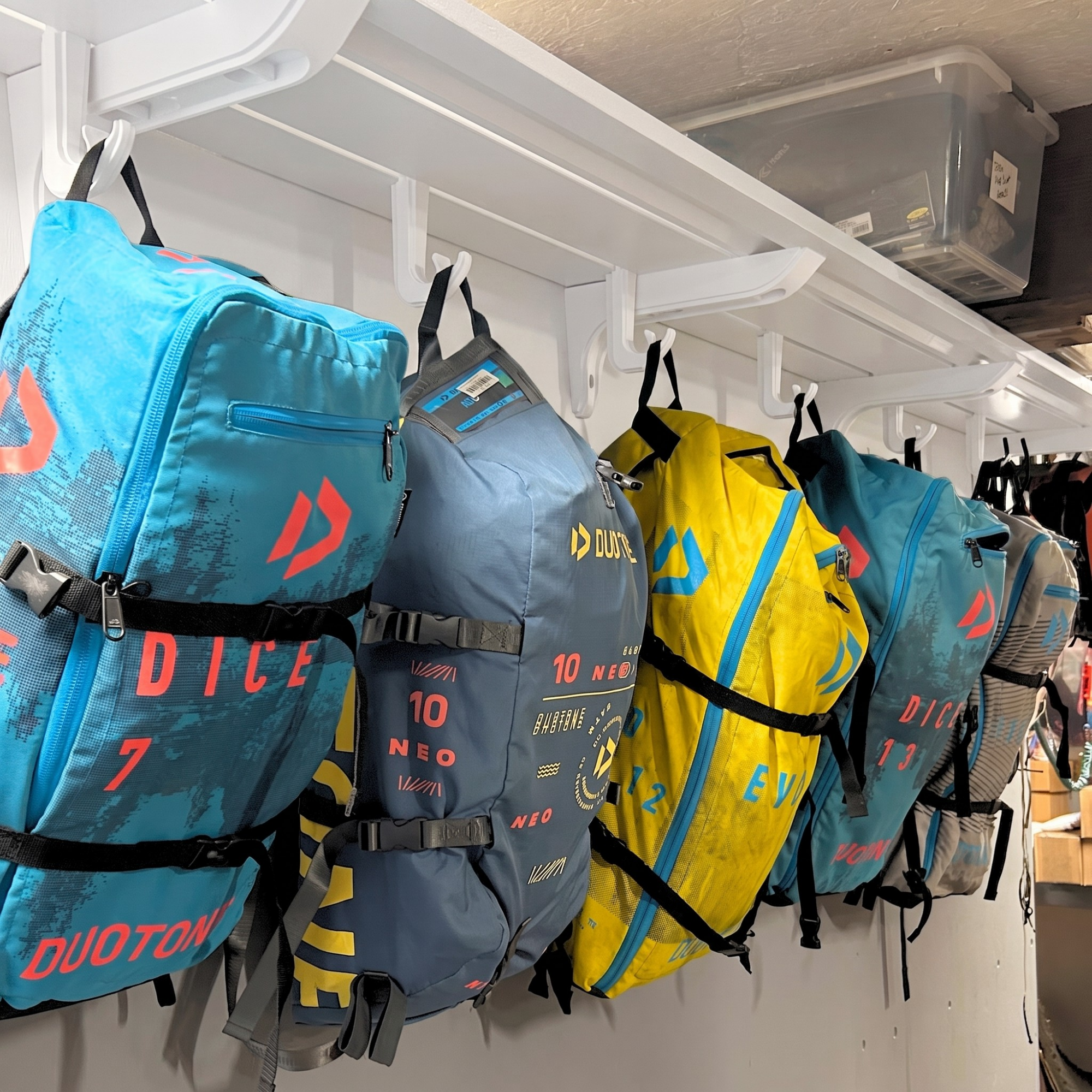 Several DUOTOY DUOTON backpacks in blue, gray, and yellow hang on white wall hooks in a storage area. The backpacks feature the red DUOTOY logo and various numbered graphics. Theee hooks could hang other types of bags as well, which would include basketball bags, baseball bags, or anything else like ocean gear