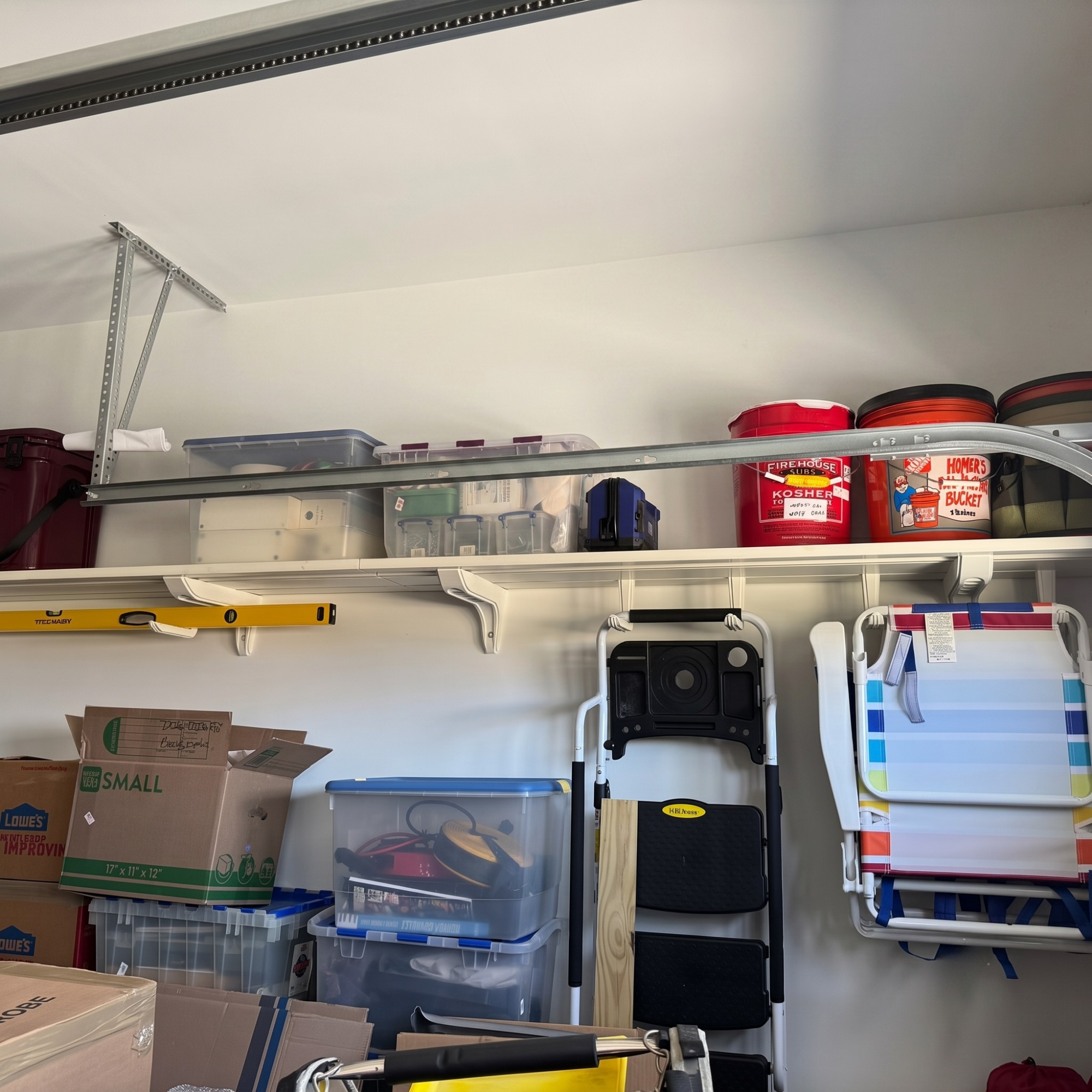 A garage storage shelf setup with white shelves mounted on a white wall, holding plastic bins, boxes, a folding chair, and red paint cans.