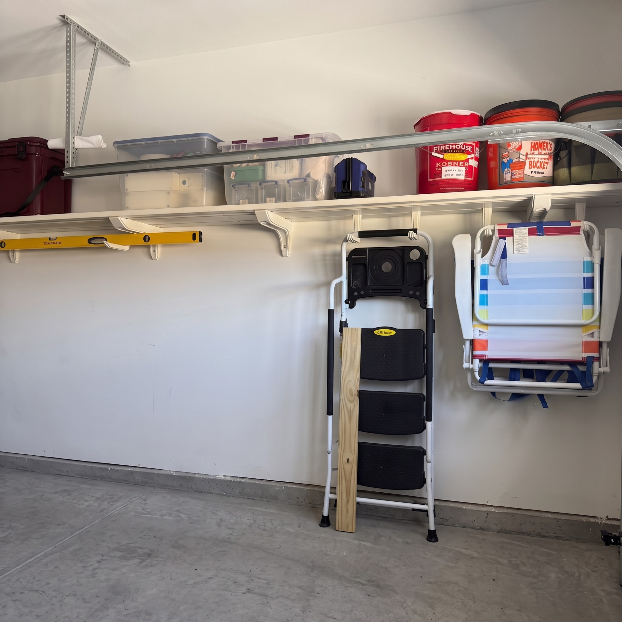 A garage storage shelf setup with white shelves mounted on a white wall, holding plastic bins, boxes, a folding chair, and red paint cans.
