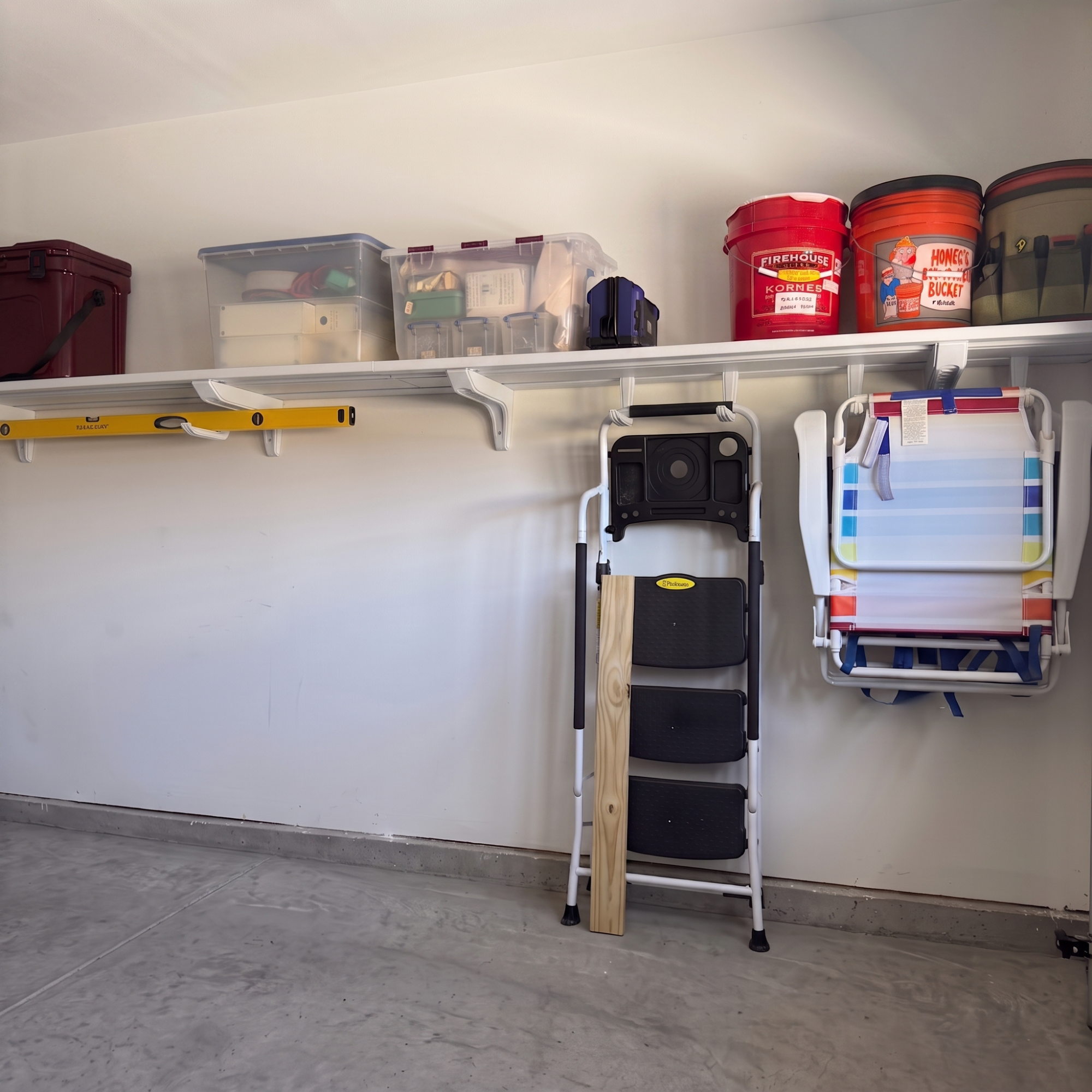 A garage storage shelf setup with white shelves mounted on a white wall, holding plastic bins, boxes, a folding chair, and red paint cans.
