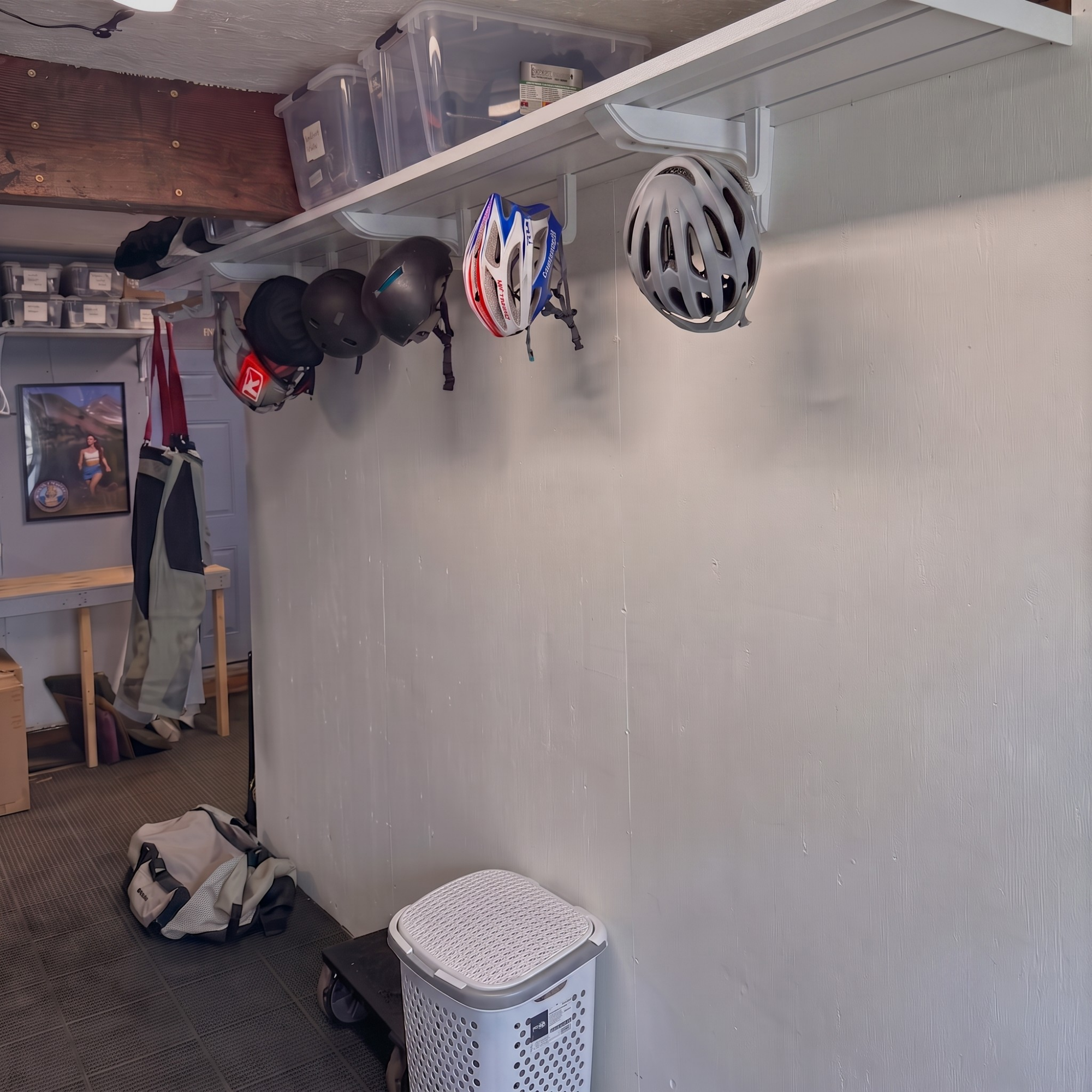 A white wall-mounted bike rack holding several bicycle helmets in a garage or storage room. A white floor bin, a laundry basket, and other room elements are visible in the background.
