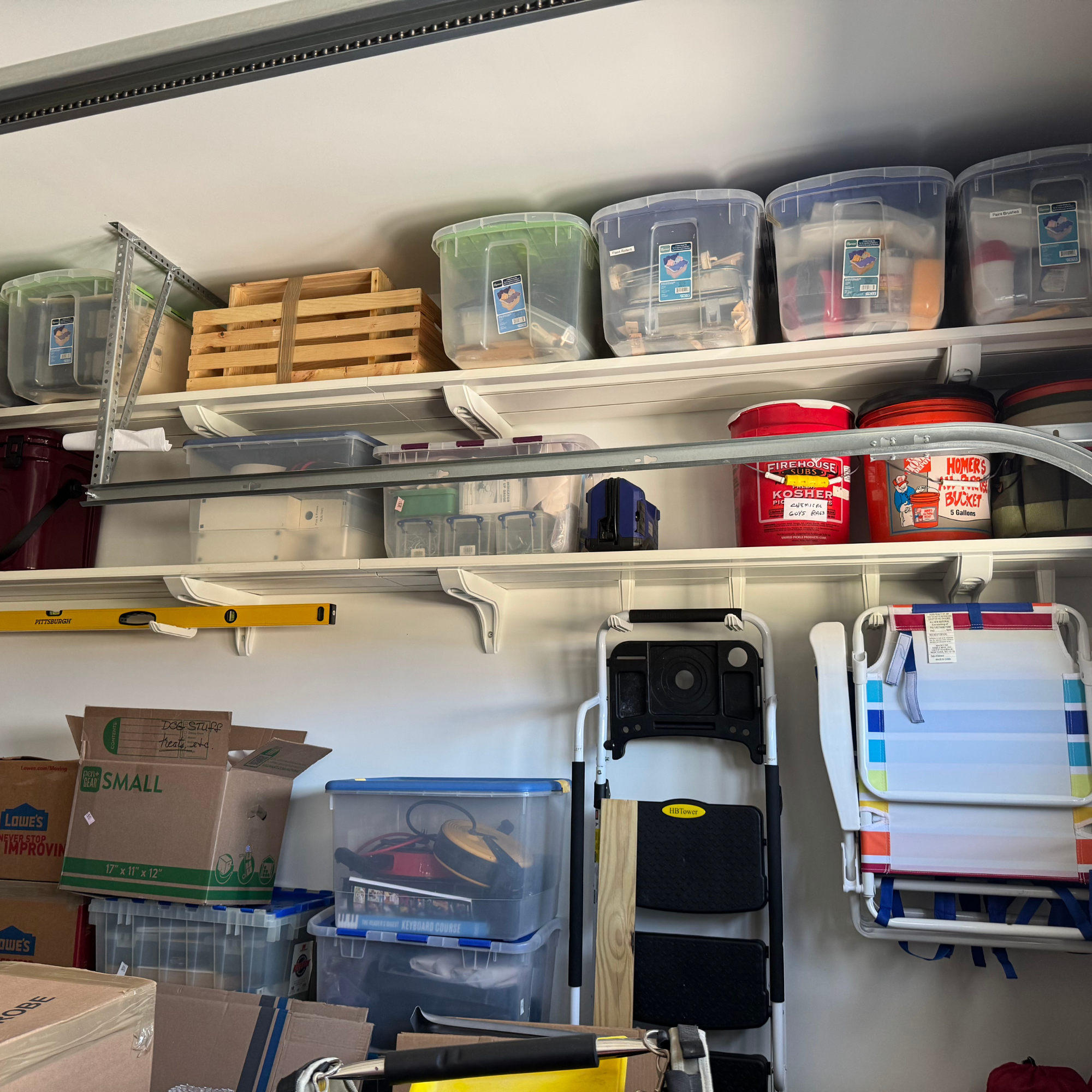 all-groups Toden heavy-duty garage storage system featuring a multi-row modular shelving configuration. White floating surfaces are mounted high on a garage wall, holding heavy plastic storage totes, toolboxes, and buckets. Integrated storage hooks and brackets are used to organize a spirit level, folding beach chairs, and a step ladder, showcasing the system's industrial load capacity and tool-free accessory integration in a high-traffic workspace.