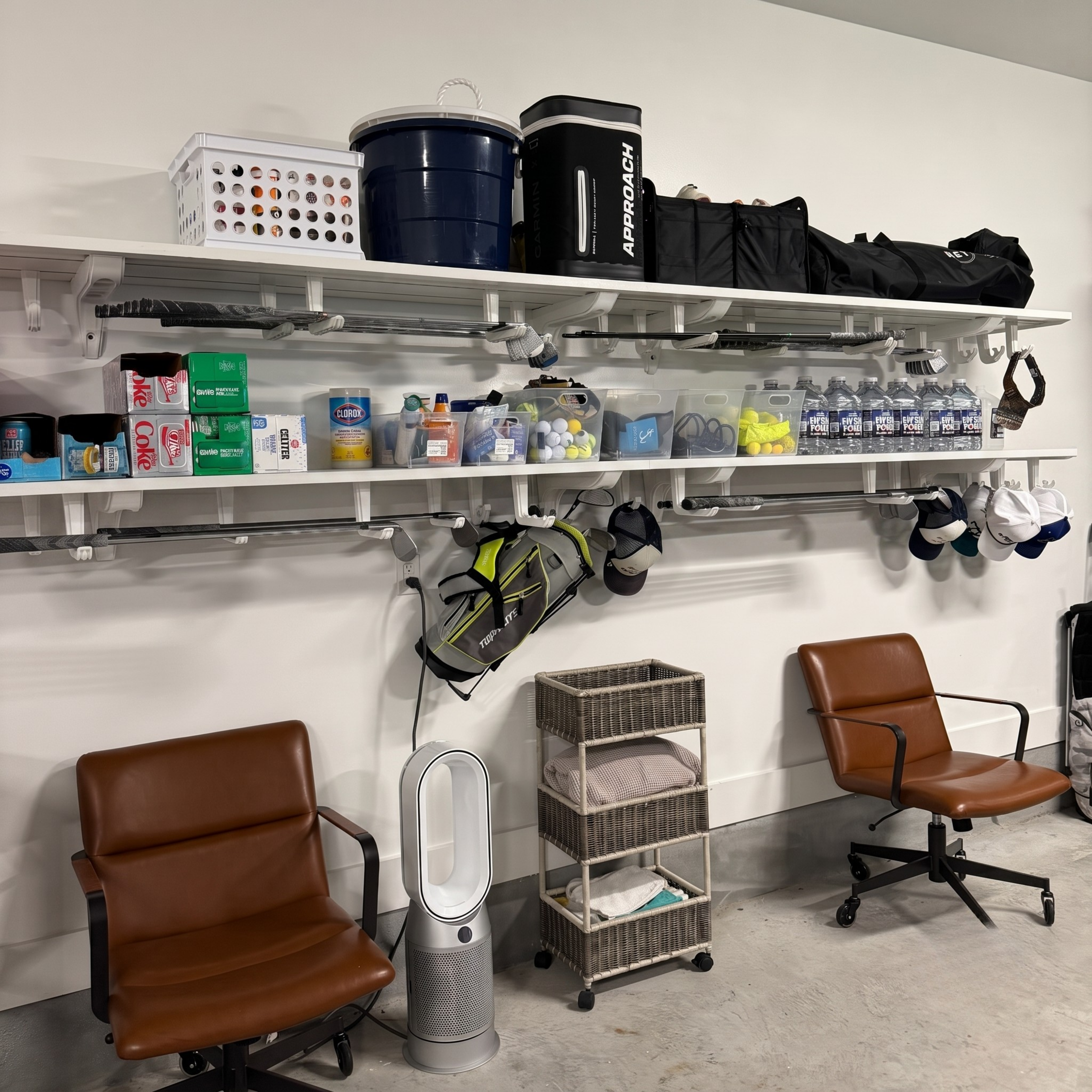A storage room with white wall-mounted shelves holding bins, coolers, bags, and assorted items. Two brown office chairs are placed in front of the shelves, along with a small rolling cart and a silver floor fan.