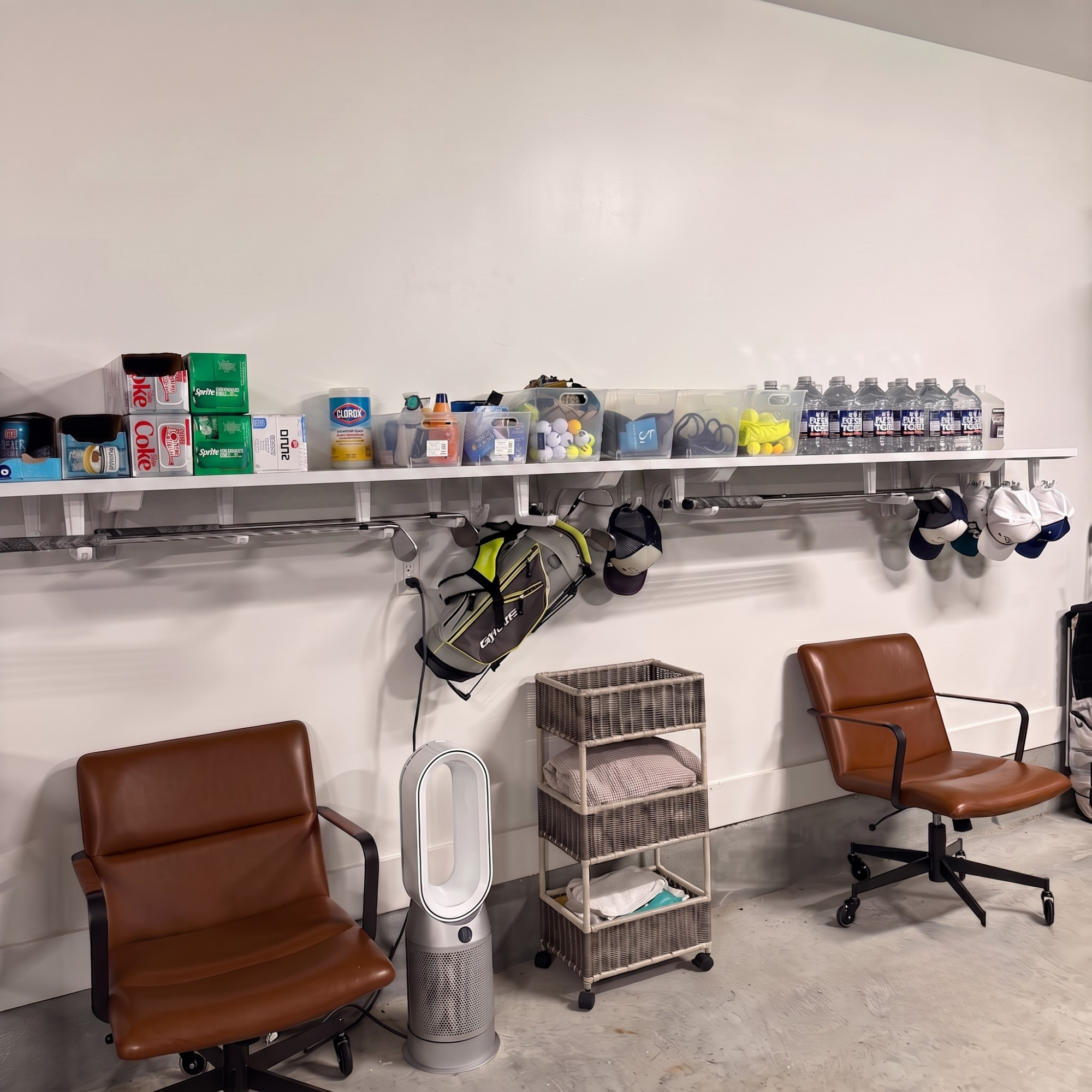 A home office or gym lounge area with two brown leather office chairs, a white wall-mounted shelf filled with various items including water bottles, boxes, and sports gear, a hanging golf bag, a small rolling storage cart, and a white tower fan on a concrete floor. Golf club storage though, for real.