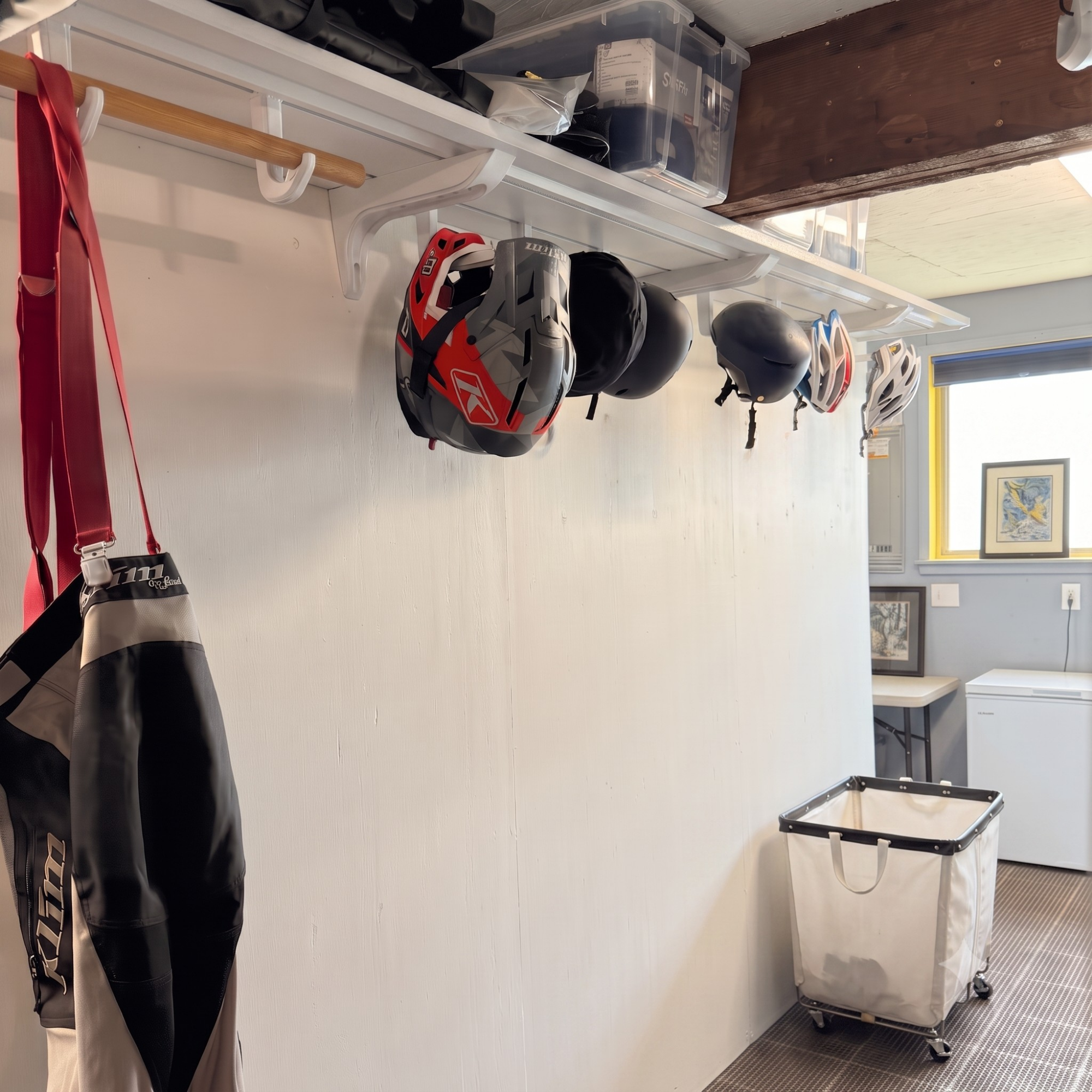 A white wall-mounted storage rack in a utility room holding several bicycle helmets, with a red-and-black bag hanging on the left and a white laundry hamper on wheels near the floor.