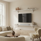 Toden white modular shelving system configured as a minimalist floating TV console and upper media shelf in a bright living room. Two long white surfaces frame a wall-mounted television, styled with books, a soundbar, and earth-toned ceramics. The setup showcases the system's sleek "no diagonal support" design and industrial-strength load capacity in a high-end, contemporary residential interior.