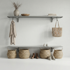 Toden modular entryway storage system in a minimalist mudroom. Two long, gray floating shelves are mounted with integrated storage hooks holding a trench coat and a woven tote bag. The bottom shelf serves as a shoe and boot station, with wicker baskets underneath for additional organization. The setup showcases the heavy-duty brackets' sleek profile and the system's "Modern Form" in a high-traffic home environment