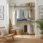 A styled closet interior with shelves and hangers holding folded and hanging clothing in soft neutral tones, alongside woven baskets, framed wall art, a chair, and a potted plant.