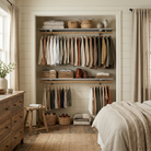 A styled bedroom interior showing a built-in closet with multiple garments hanging on racks, folded textiles, baskets, and a bed with a chunky knit blanket in the foreground.