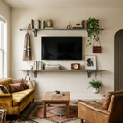Toden modular shelving system used as a modern entertainment center and floating TV console in a curated, organic modern living room. Two long gray surfaces are mounted on a neutral wall, framing a flat-screen television. The shelves are styled with books, plants, and art, while integrated snap-fit hooks are used to hang a scarf, a leather bag, and a potted plant. The setup showcases the "no diagonal support" design and the system's ability to blend industrial strength with high-end residential decor.