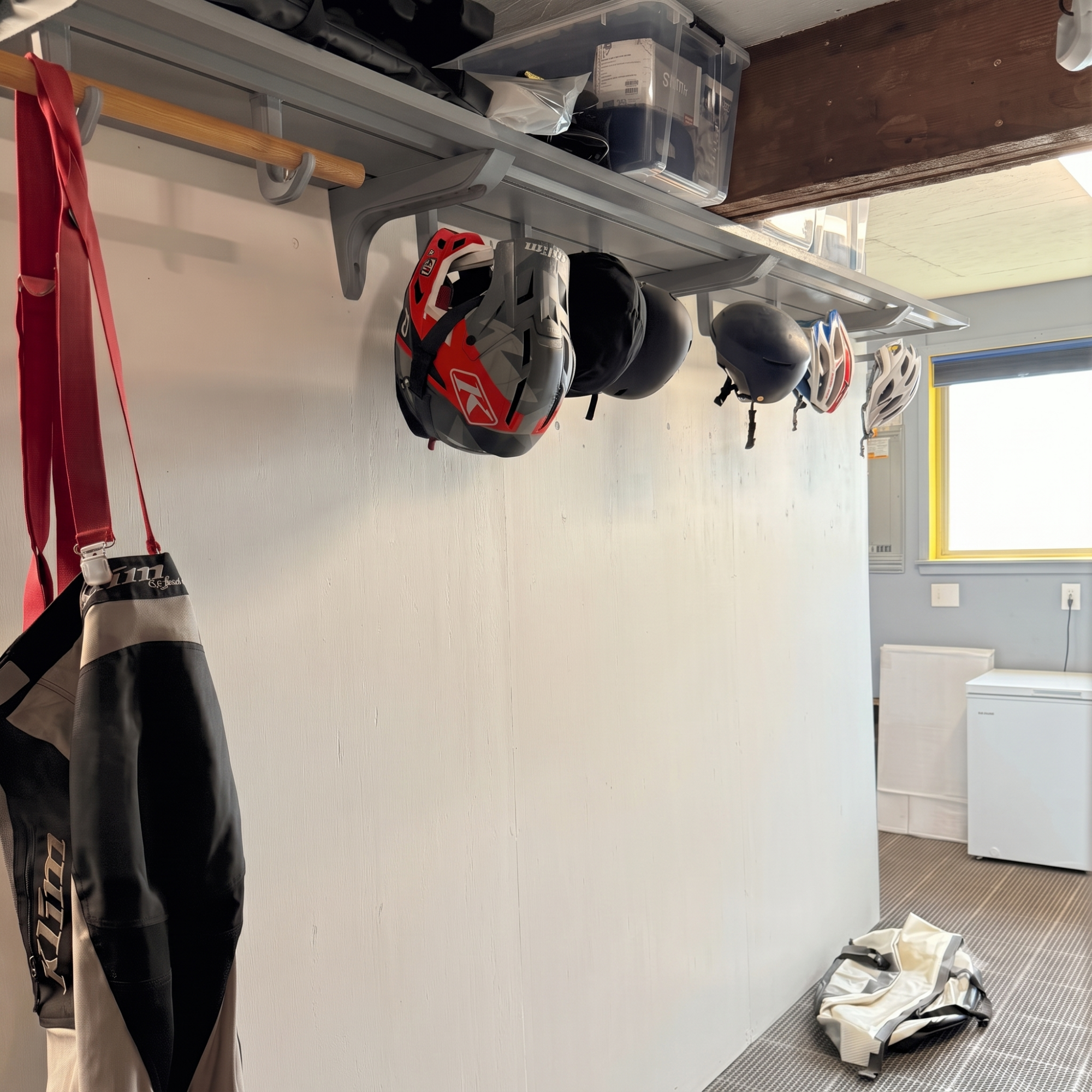 A wall-mounted storage rack in a garage or workshop holding several bicycle helmets, with a red-and-black bag hanging on the left and storage bins on the shelf.
