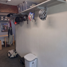 A gray wall-mounted bike rack holding several bicycle helmets in a garage or storage room. A white laundry hamper sits on the floor, and other room items like a table, storage bins, and a backpack are visible in the background.