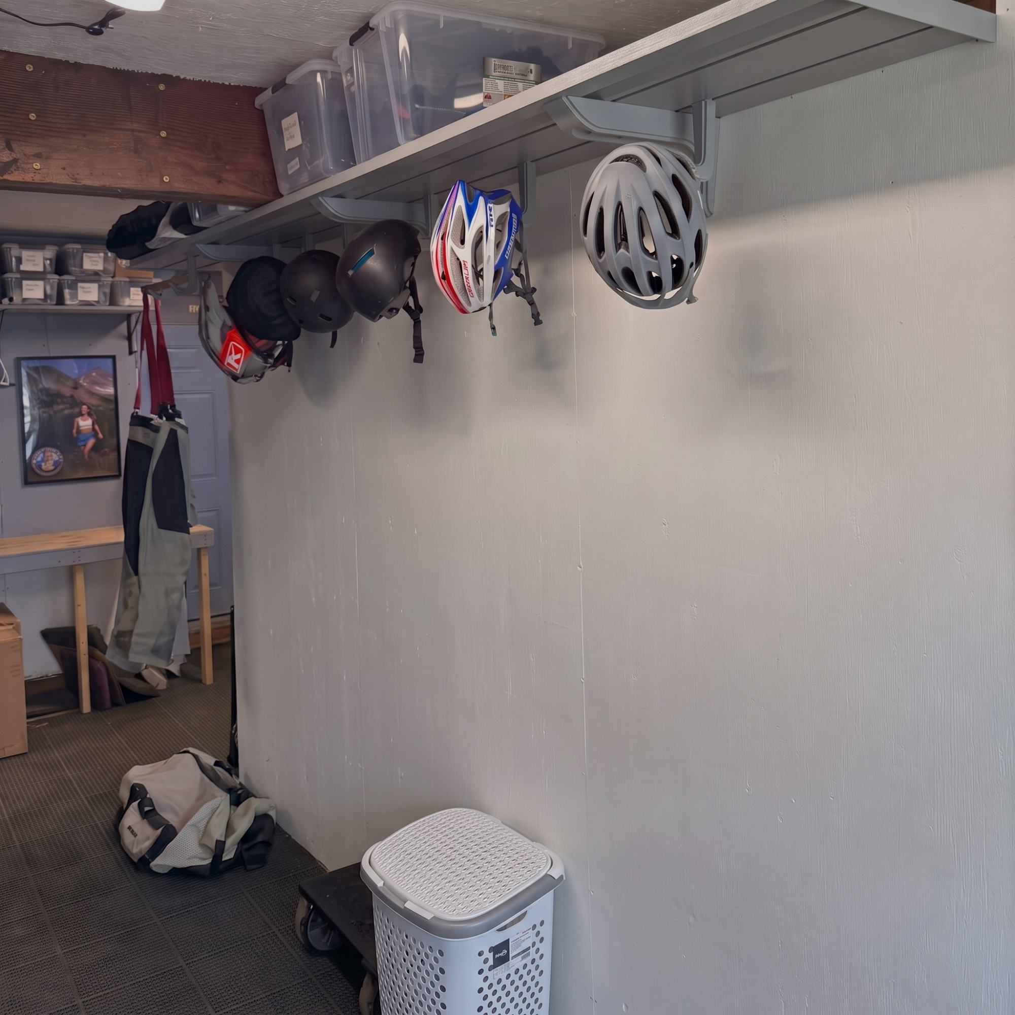 A gray wall-mounted bike rack holding several bicycle helmets in a garage or storage room. A white laundry hamper sits on the floor, and other room items like a table, storage bins, and a backpack are visible in the background.