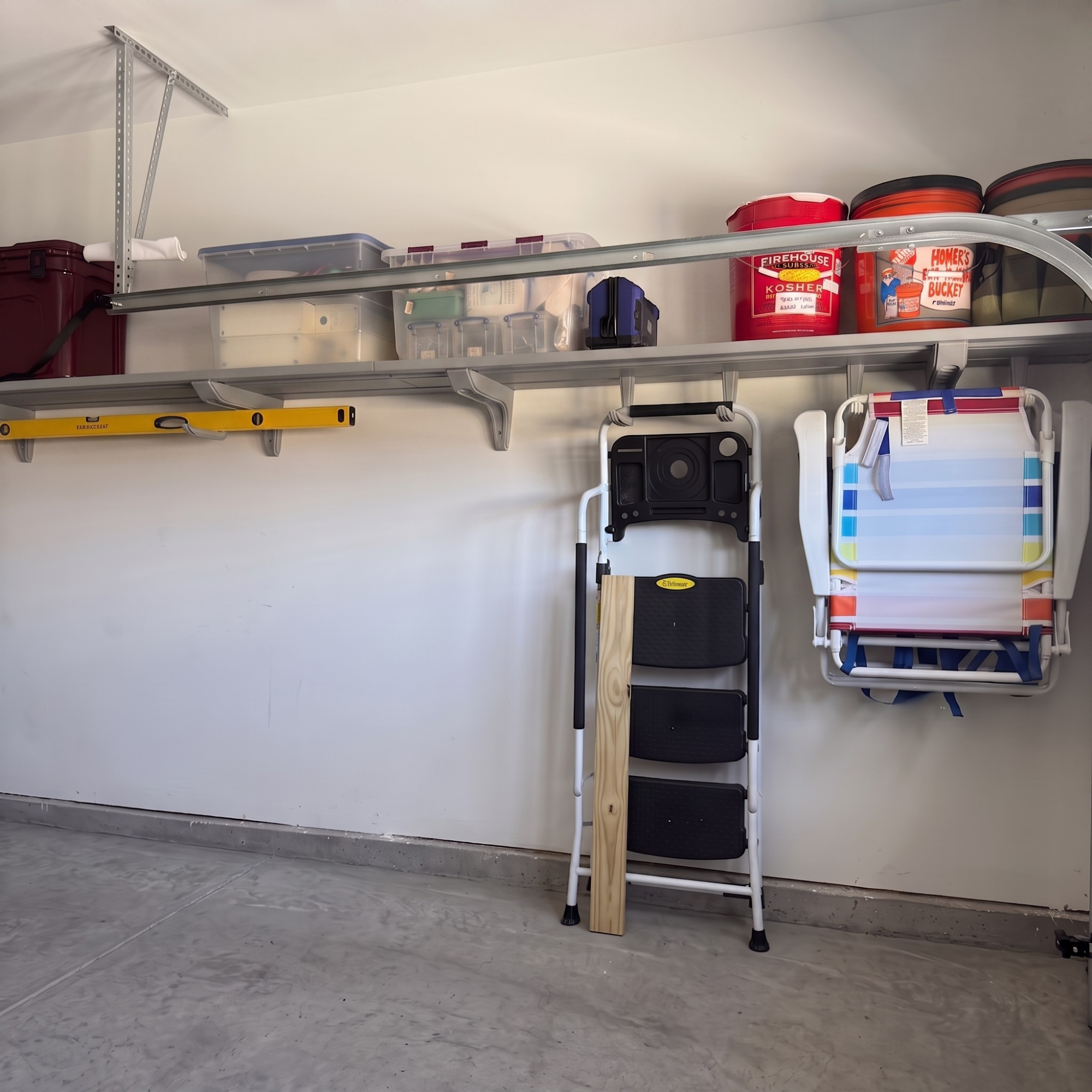 A metal garage wall-mounted storage shelf system holding various household items, including plastic bins, buckets, a yellow level, a folding step ladder, and a white folding chair, against a white garage wall.