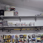 A storage area with gray shelves holding clear plastic organizer bins, fishing rods, buckets, and a pegboard below filled with packaged fishing supplies and tackle.