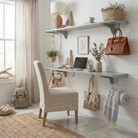 Toden modular shelving system used as a minimalist floating desk and office organizer in a bright home workspace. Two long gray surfaces are mounted on a white shiplap wall; the lower panel serves as a sleek desk for a laptop, while the upper shelf provides decorative storage. Integrated snap-fit hooks are used to hang a designer leather bag, headphones, and a tote, demonstrating the system's "Built-in Brilliance" for cable-free, organized living.