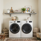 Toden gray modular shelving system installed as a laundry room organizer above a white front-load washer and dryer. The long-run floating shelf is styled with laundry supplies, plants, and art, highlighting the "no-diagonal support" design that keeps the space open and minimalist. The industrial-strength brackets provide a sleek, low-profile look for home organization.