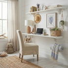 Toden white modular shelving system configured as a minimalist floating desk and home office workstation. The setup features a long white surface panel acting as a sleek desk for a laptop, with a matching upper shelf for decor and books. Integrated white storage hooks are used to hang a sun hat, headphones, and a blanket, showcasing the system's "Built-in Brilliance" and no-diagonal support design on a white shiplap wall.
