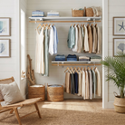 A styled closet interior with open shelving and hanging racks filled with folded and hung clothing in neutral and blue tones, alongside woven baskets, framed wall art, a rattan chair, and a potted plant.