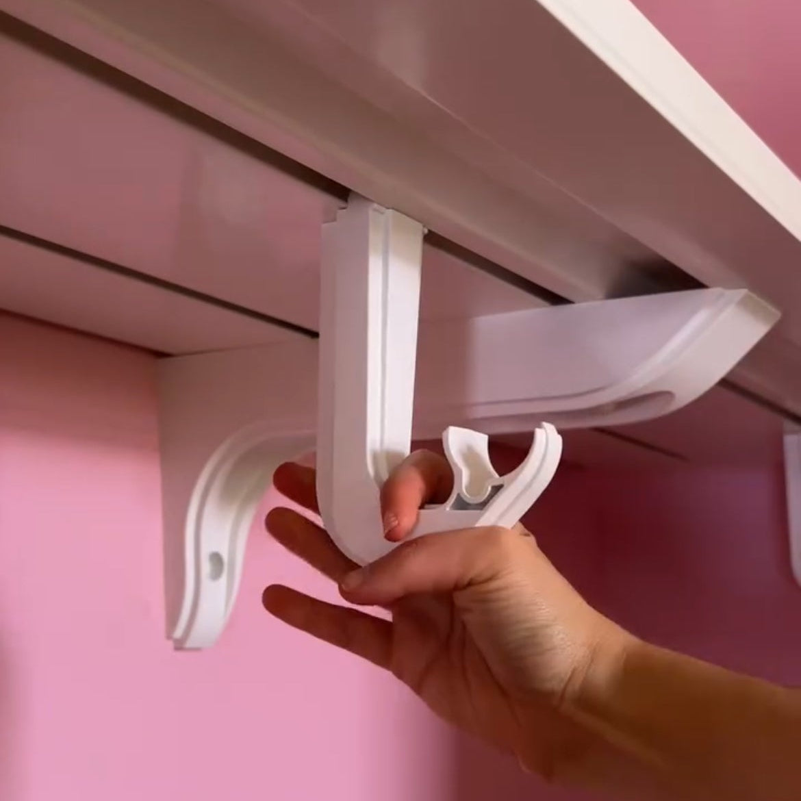 A hand holding a white plastic shelf bracket against a pink wall and white shelf frame.