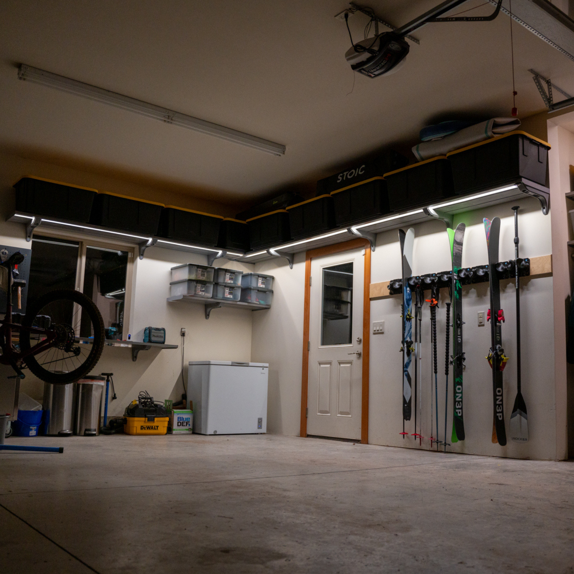 A garage interior with wall-mounted storage cubbies, a ski rack holding skis and snowboards, shelving, a chest freezer, and a bicycle wheel visible on the left.
