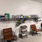 A storage room or lounge area with two brown leather office chairs, a metal shelf holding assorted bottles, boxes, and bags, and a small rolling storage cart against a white wall.