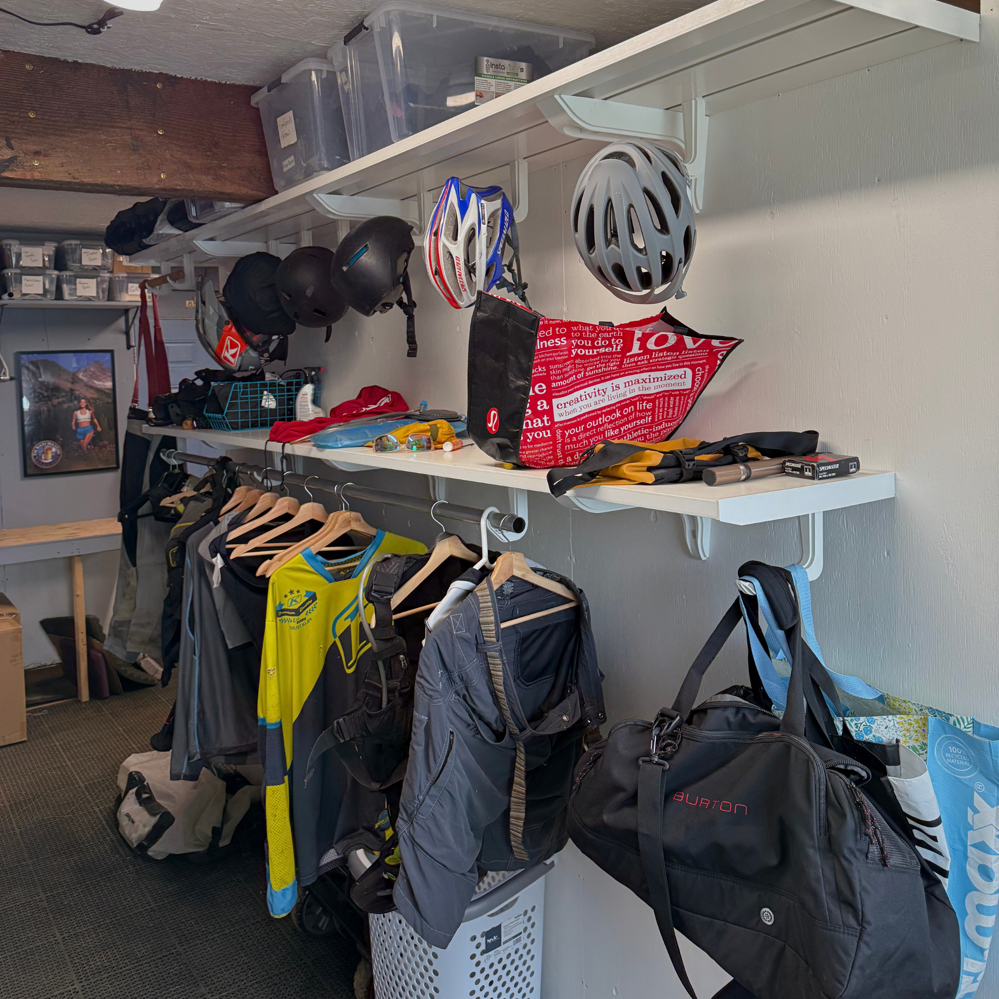all-groups Toden white modular gear room system featuring a double-row shelving layout. The upper shelf uses integrated snap-fit hooks to organize multiple helmets and gear bags. The lower shelf includes a heavy-duty closet rod for hanging jerseys, jackets, and tactical vests. The setup demonstrates how the system maximizes vertical wall space for bulky athletic gear and apparel in a garage or mudroom setting.