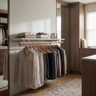 A styled walk-in closet interior with a wooden hanging rod holding multiple shirts on wooden hangers, a floating shelf with folded textiles and books, and warm wood cabinetry in the background.