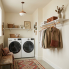 Toden white modular laundry room system featuring a double-wall configuration. One white floating shelf is mounted above the washer and dryer for detergent and basket storage, while a second shelf includes an integrated wooden closet rod for air-drying clothes and organizing outerwear. The setup highlights the industrial-strength brackets and sleek, no-diagonal support design in a warm, modern residential utility space.
