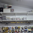 Toden heavy-duty garage shelving system used as a fishing gear organization station. Two white modular shelves are mounted above a pegboard wall, holding heavy buckets, a cooler, and dozens of plastic tackle organizers. Integrated storage hooks are used as a horizontal fishing rod rack, securing multiple rods along the underside of the shelf. The setup highlights the system's massive load capacity and its ability to organize specialized equipment in a workspace or garage.