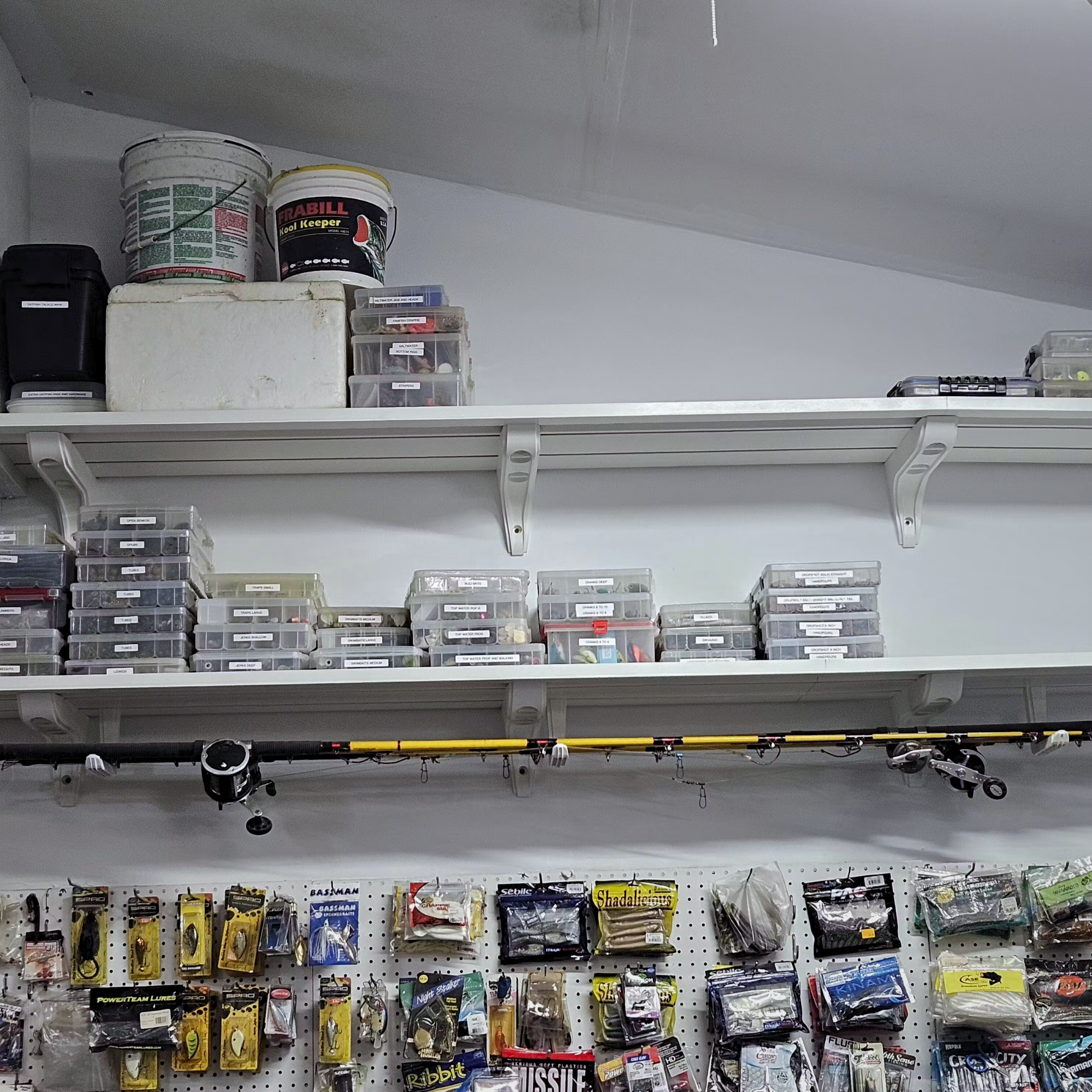 Toden heavy-duty garage shelving system used as a fishing gear organization station. Two white modular shelves are mounted above a pegboard wall, holding heavy buckets, a cooler, and dozens of plastic tackle organizers. Integrated storage hooks are used as a horizontal fishing rod rack, securing multiple rods along the underside of the shelf. The setup highlights the system's massive load capacity and its ability to organize specialized equipment in a workspace or garage.