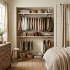 A styled bedroom interior showing a built-in closet with shelves and hangers filled with neutral-toned clothing, alongside a bed, dresser, stool, baskets, and curtains.